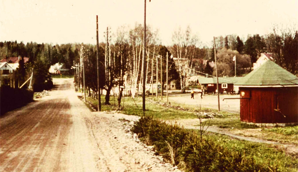 1960-luvun alussa alas kohti keskustaa vievää Thurmanin puistotietä levennettiin. Siellä, mihin tie aikaisemmin päättyi, oli vielä silloin pumppuhuone josta ihmiset saivat hakea vettä. Nykyään sen paikalla on kaupungintalo. Urheilukenttä pumppuhuoneen alapuolella oli nuorison suosima kokoontumispaikka. Siellä oli mm. kelkkamäki talviaikaan. 1960-luvulla, kun asukasmäärä kasvoi parilla tuhannella muutamassa vuodessa ja Kauniainen kuhisi lapsia, saatiin sinne myös jääkiekkokenttä. Sillä paikalla on nykyään kuvanveistäjä Heikki Häiväojan tekemä sotainvalidien muistomerkki, joka vihittiin käyttöön 1981. Kuvassa tie jatkuu kaartuen yli rautatien. I början av 1960-talet hade Thurmansallén ner mot centrum breddats. I den gamla sluttningen stod då ännu ett pumphus med vatten för allmänheten. I dag står stadshuset på dess plats. Sportplanen nedanför pumphuset var en populär samlingsplats för ungdomar med bland annat en kälkbacke vintertid. På 1960-talet då invånarantalet steg med ett par tusen på några år och Grankulla kryllade av barn fick planen en ishockeyrink. Det är där krigsinvalidmonumentet står i dag, gjort av skulptören Heikki Häiväoja och invigt 1981. På bilden fortsätter vägen i en kurva över järnvägen. 